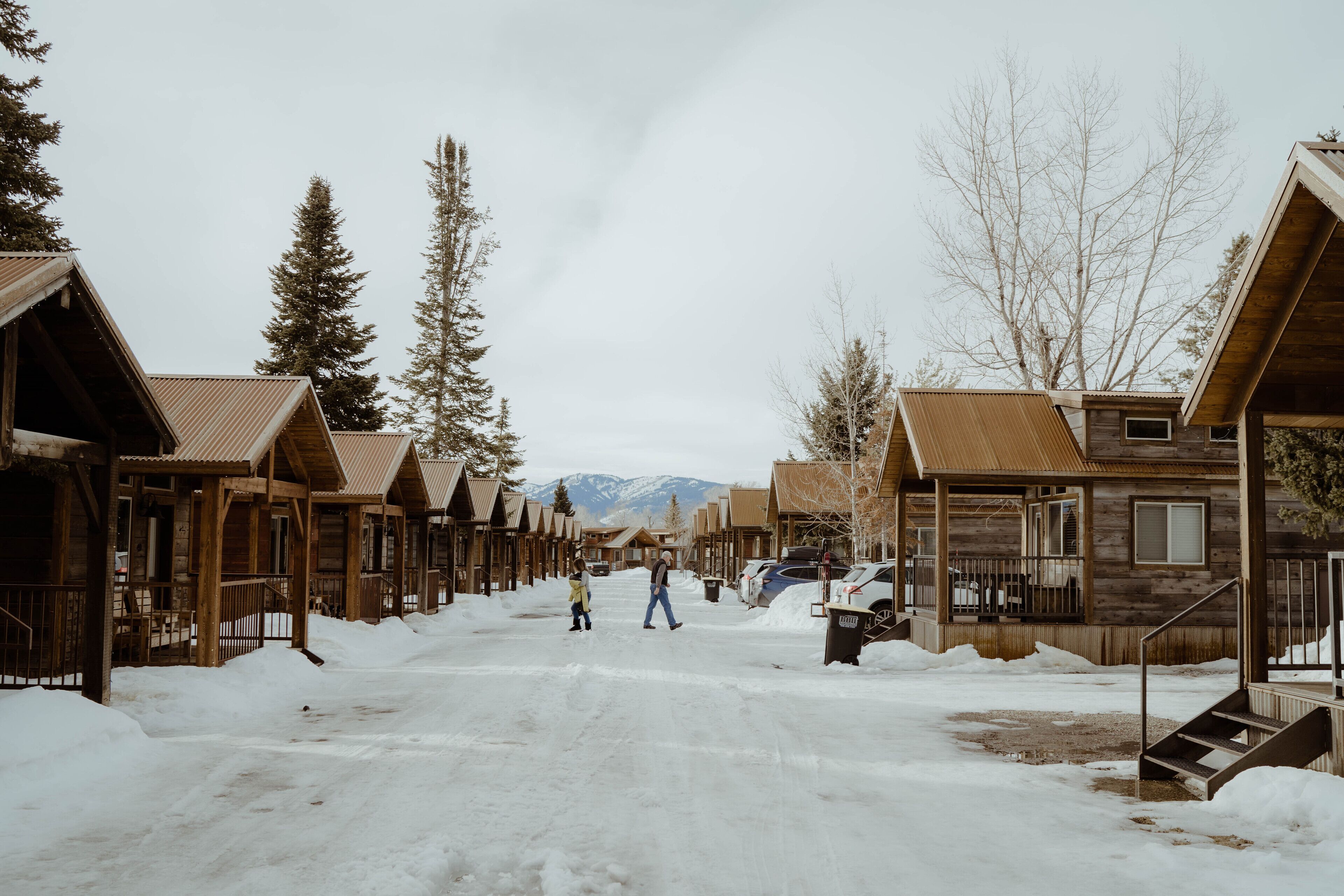 A snow-covered street lined with wooden cabins, a person walking, and a child.
