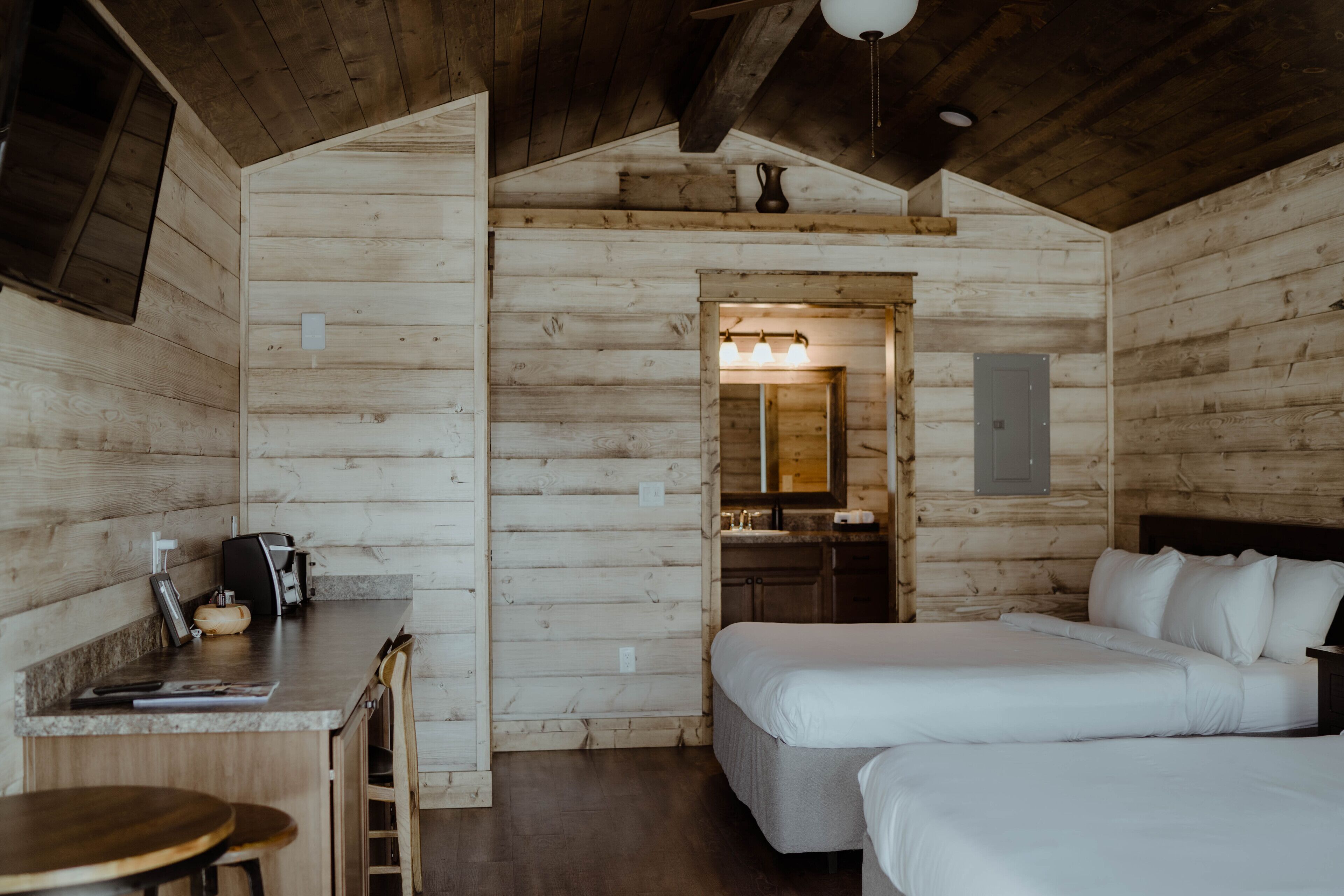 A wooden cabin interior with a kitchenette, two beds, a dining area, and a bathroom.