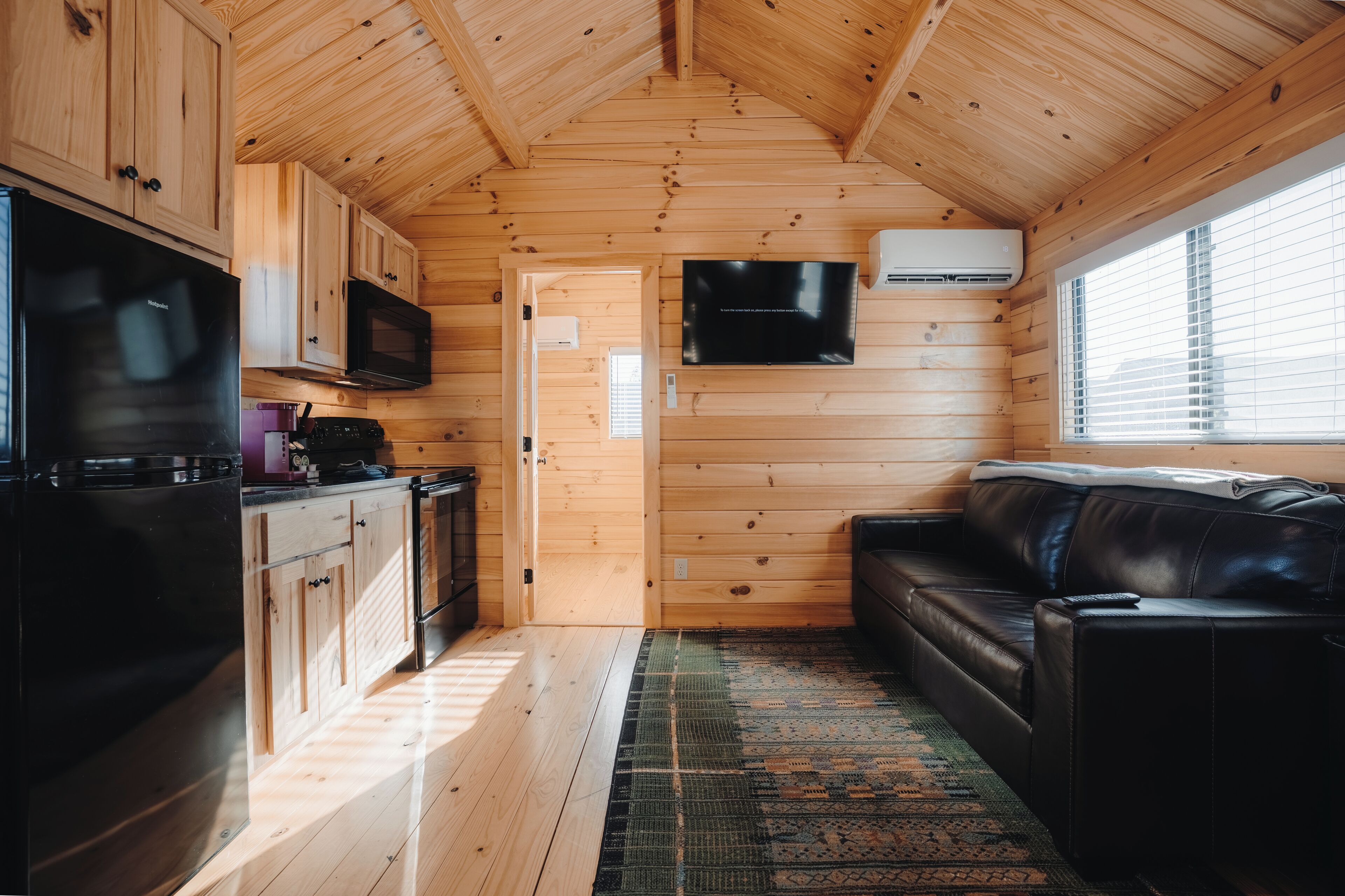 Une chambre de style cabine, de taille réduite, comprenant un coin cuisine, un canapé en cuir noir et un téléviseur fixé au mur.