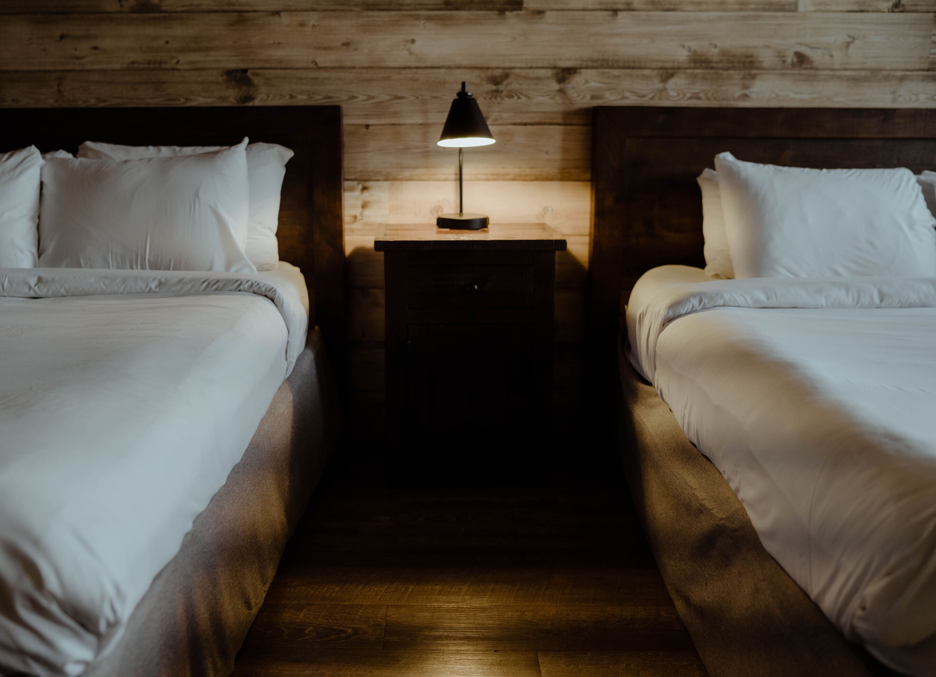 A room with two beds, a wooden headboard, a nightstand with a lamp, and a wooden floor.