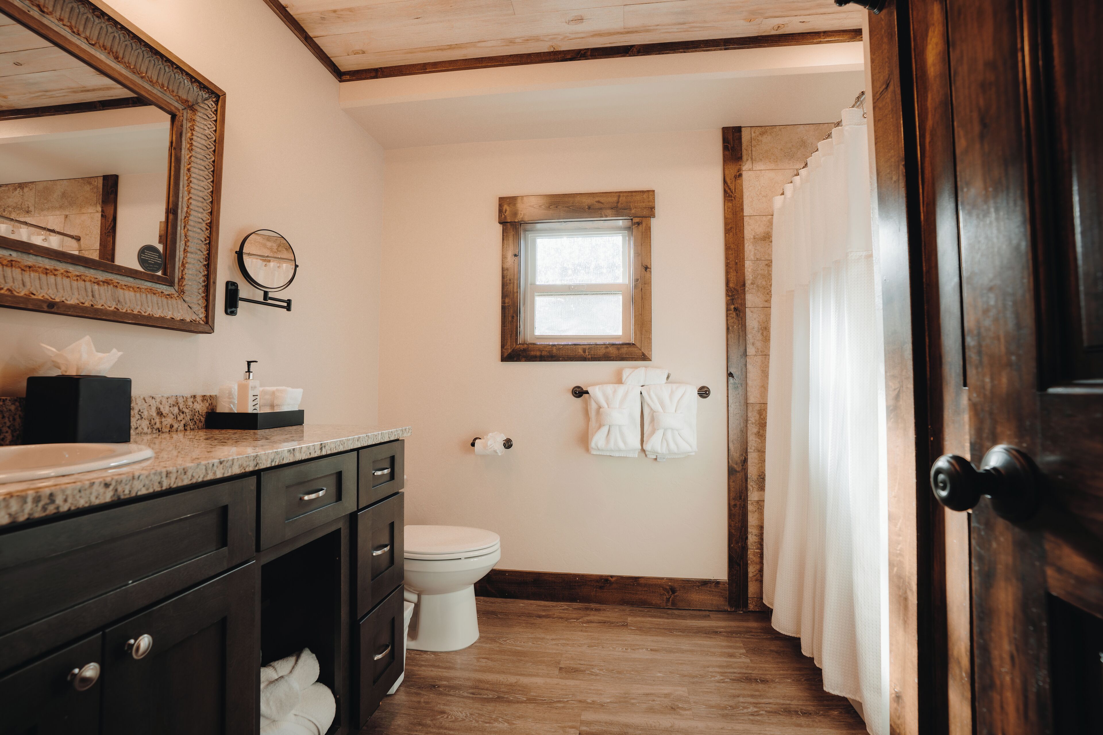 A bathroom with a wooden vanity, a large mirror, a small window, and a door leading to a shower area.