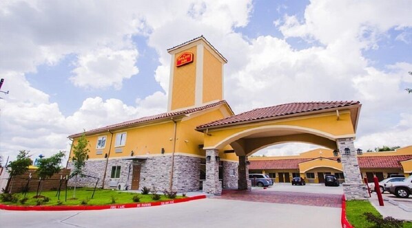 A hotel with a yellow facade, a stone base, and a red-tiled roof. There is a sign on the tower and a covered entrance.