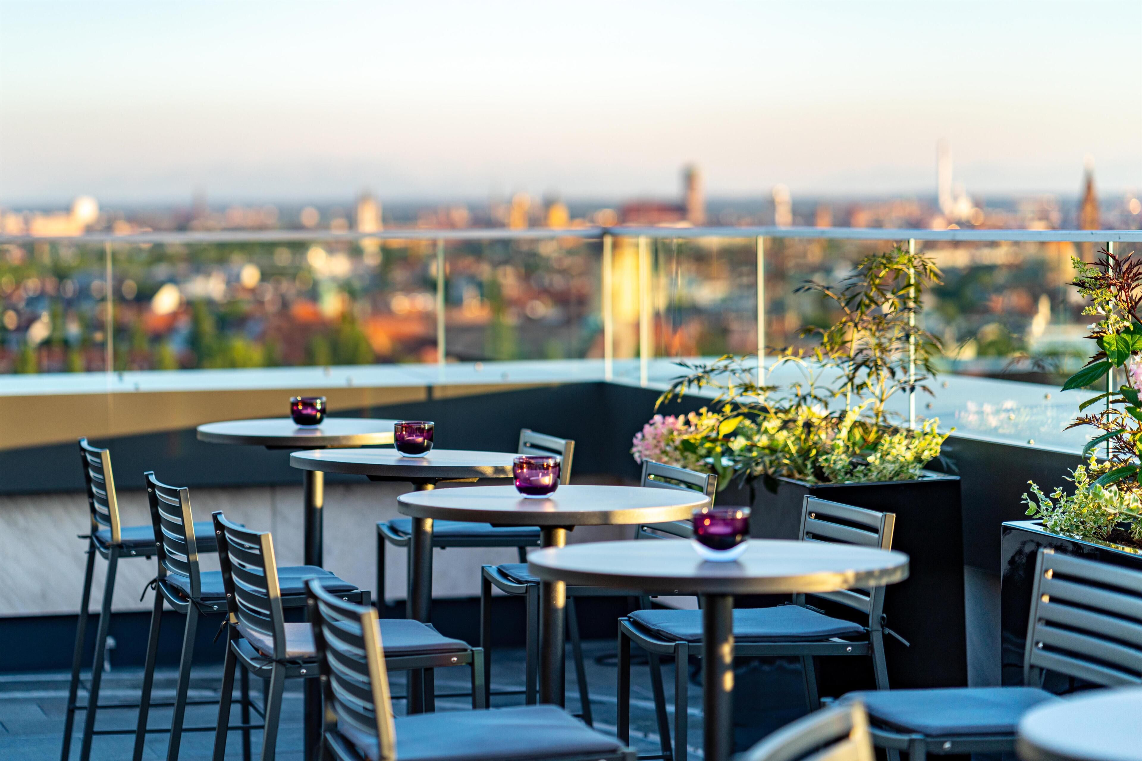 Una terraza en la azotea con mesas y sillas, vistas a la ciudad y plantas en macetas.