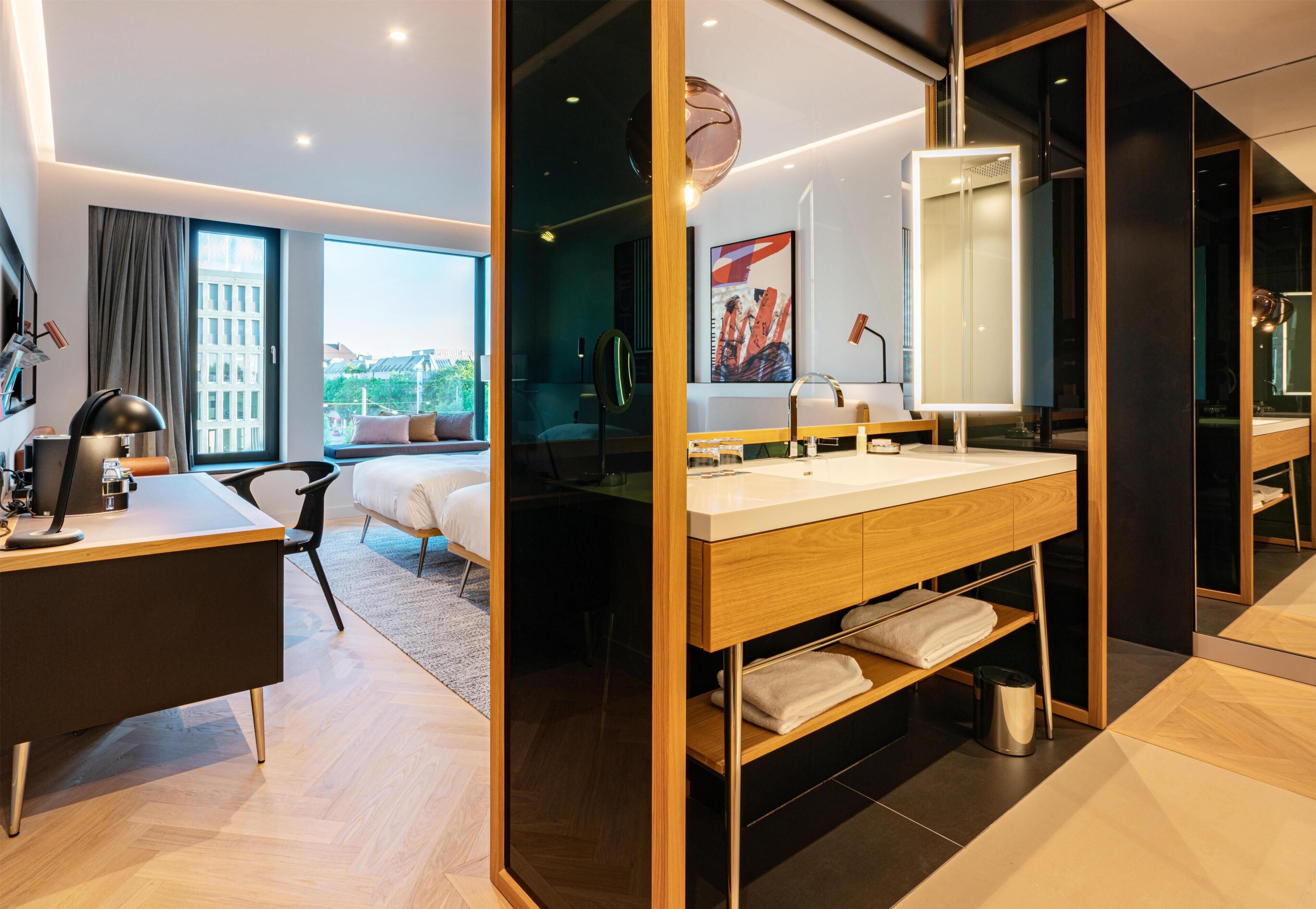 Una habitación de hotel moderna con una cama grande, un escritorio, un baño con tocador de madera y un ventanal con vistas a la ciudad.