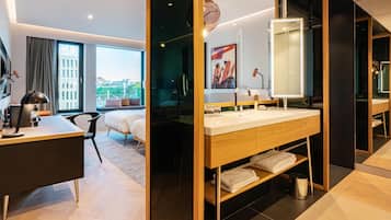 Una habitación de hotel moderna con una cama grande, un escritorio, un baño con tocador de madera y un ventanal con vistas a la ciudad.