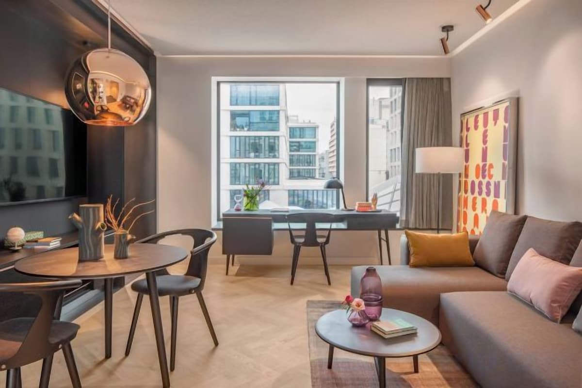 Una sala de estar moderna con sofá, mesa de centro y comedor. Hay un ventanal con vista a edificios.