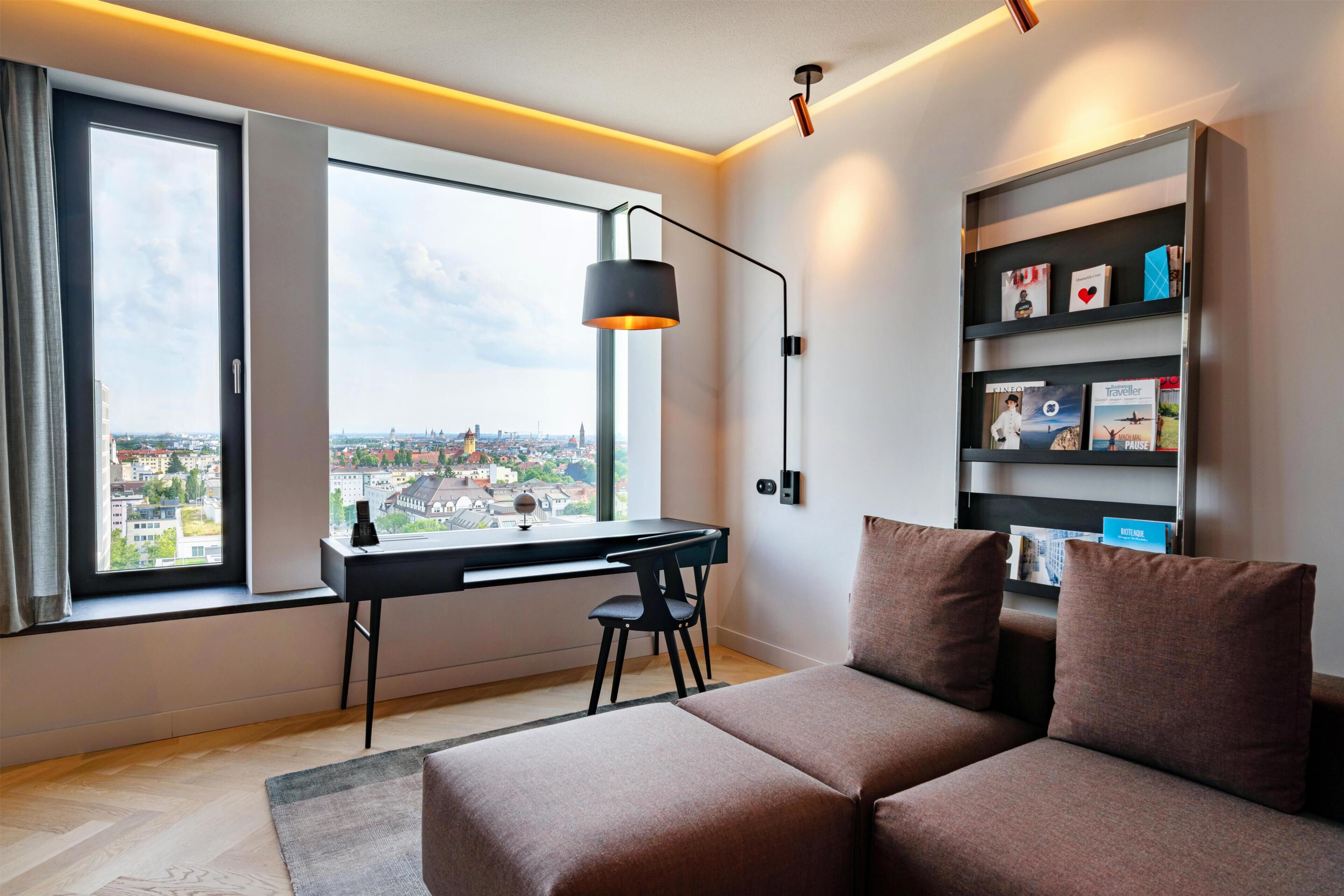 A modern room with a desk, chair, and sofa, a bookshelf, and a large window with a city view.