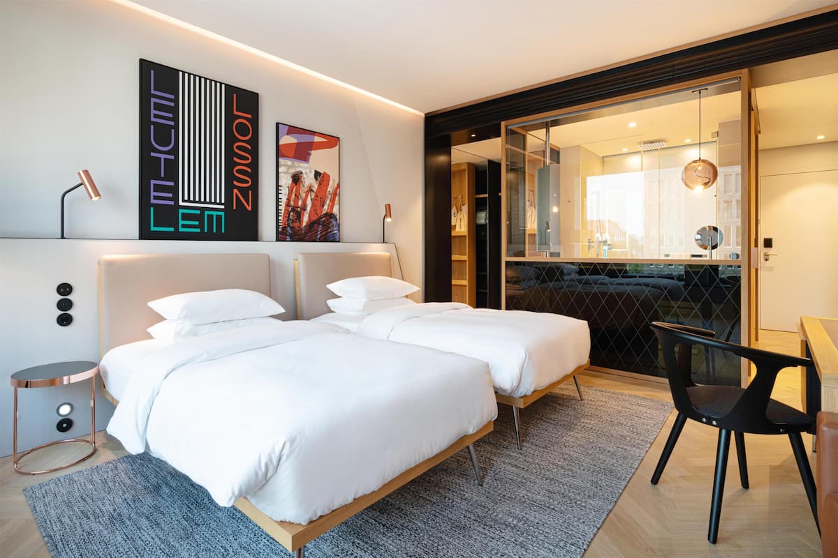 Una habitación de hotel moderna con una cama doble, una mesita auxiliar pequeña, una silla y un ventanal grande.