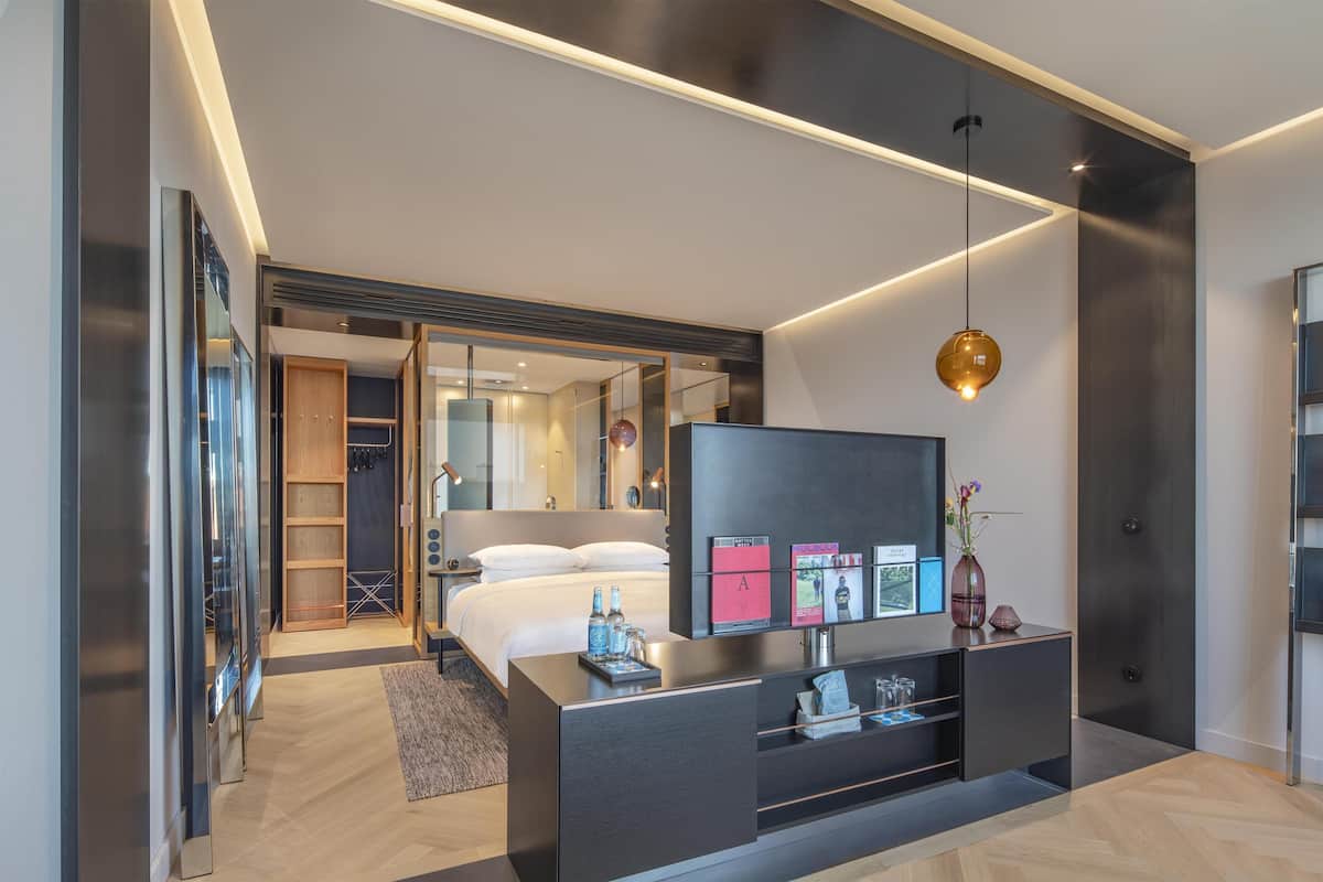 Una habitación de hotel moderna con una cama, un televisor de pantalla plana, una mesita de consola con objetos y un pasillo que conduce a un baño.