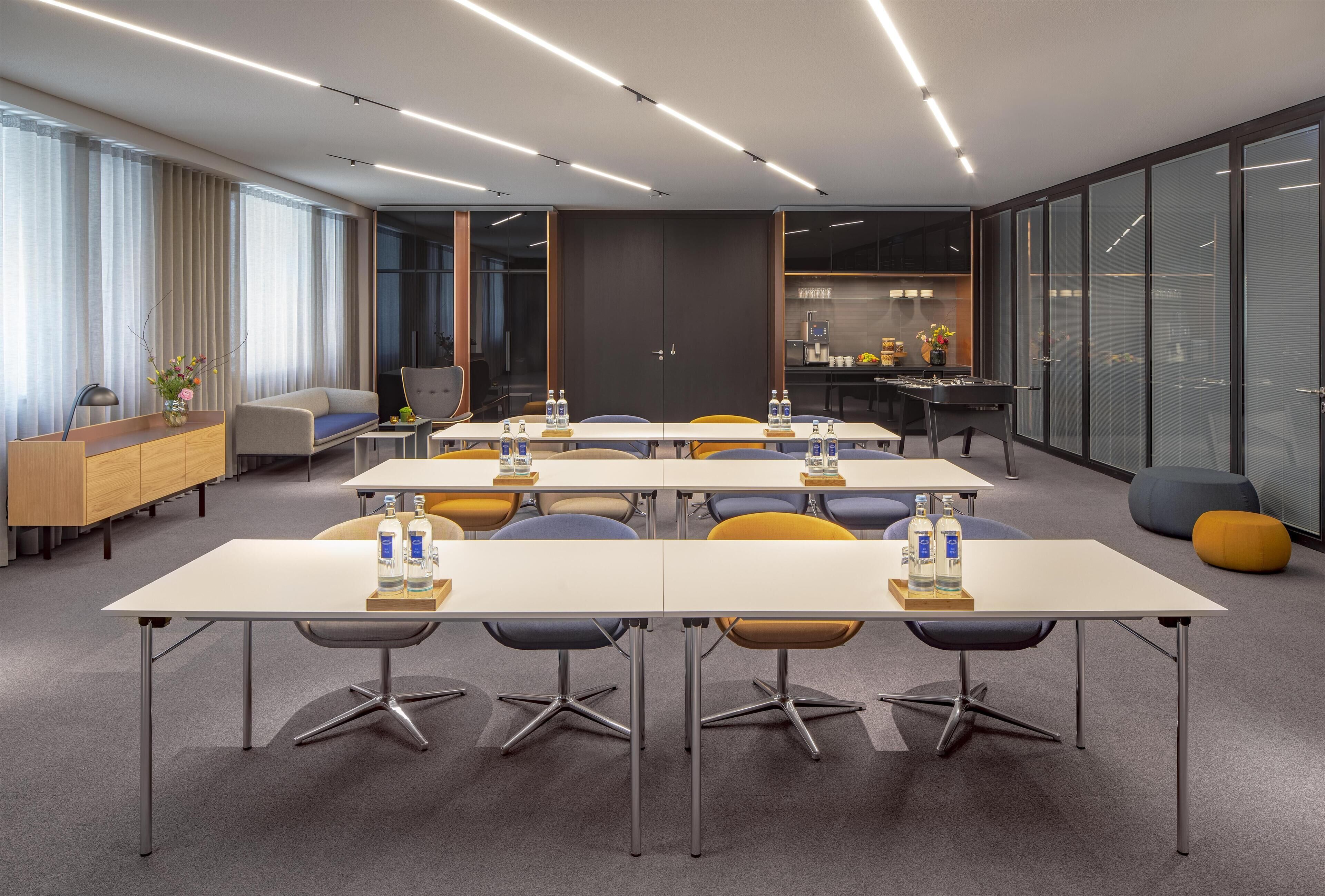 A modern conference room with multiple tables, chairs, and a kitchenette area.