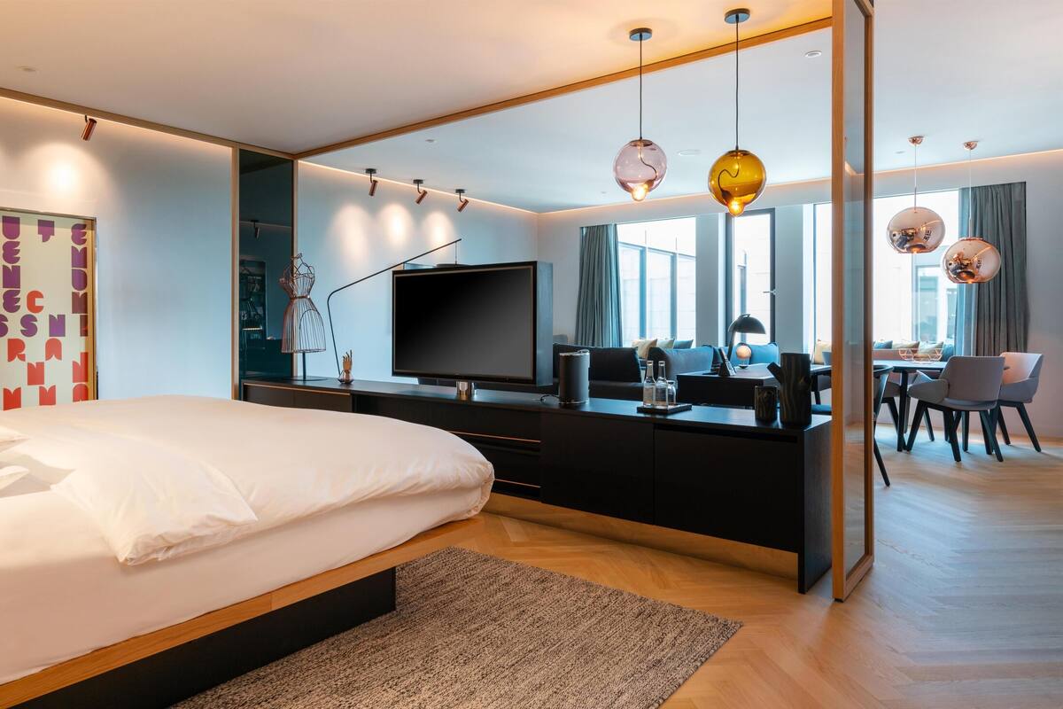 Una habitación de hotel moderna con una cama grande, un televisor de pantalla plana, un escritorio con silla y un comedor con mesa y sillas.