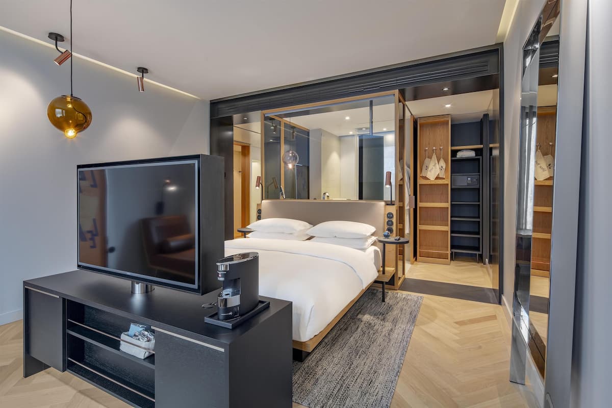 Una habitación de hotel moderna con televisión de pantalla plana, cafetera, cama con ropa de cama blanca, un espejo grande y un vestidor.