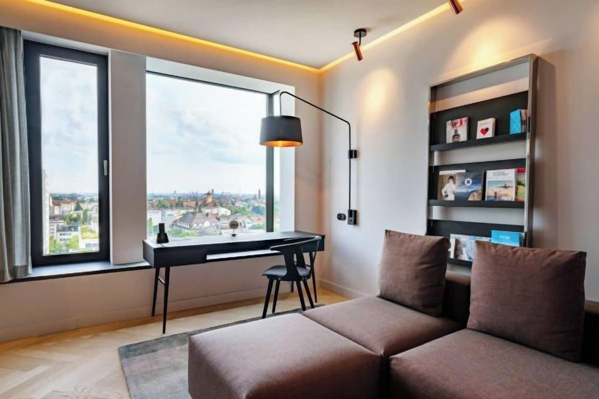 Una habitación moderna con escritorio, sofá, estantería y un amplio ventanal con vista a la ciudad.