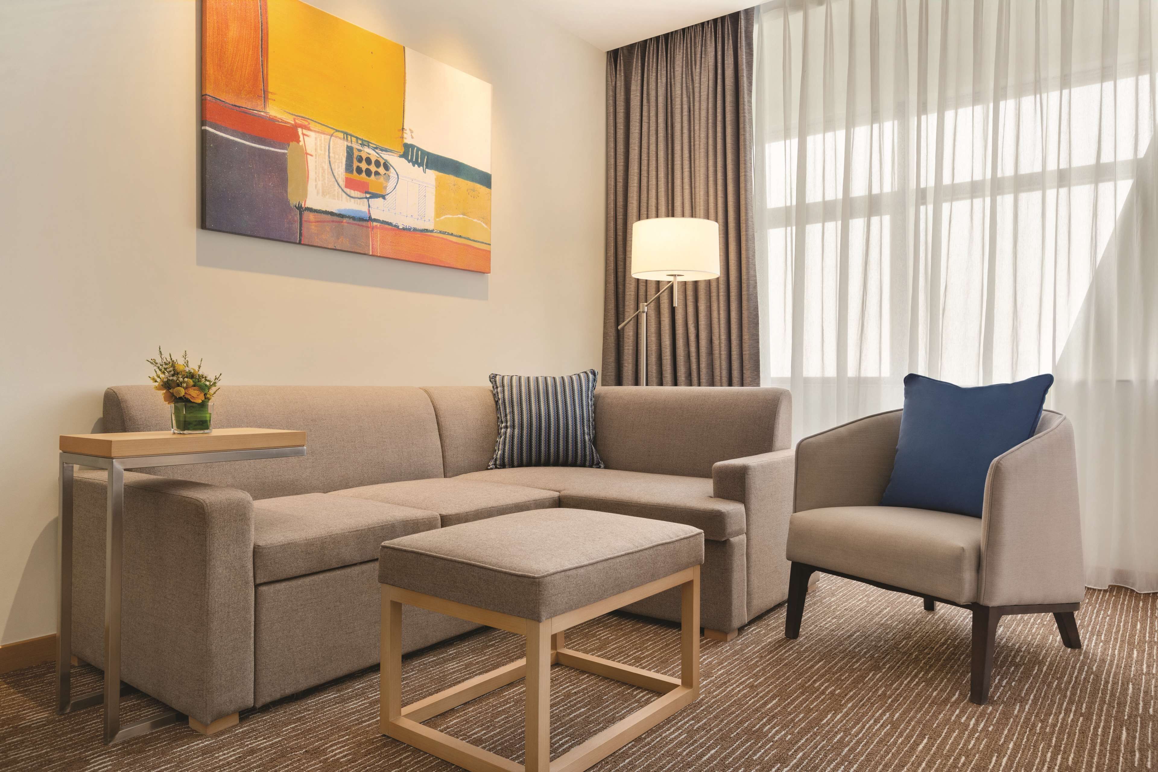 Habitación de hotel con sofá modular, un sillón individual, una mesita, una lámpara y un cuadro en la pared.
