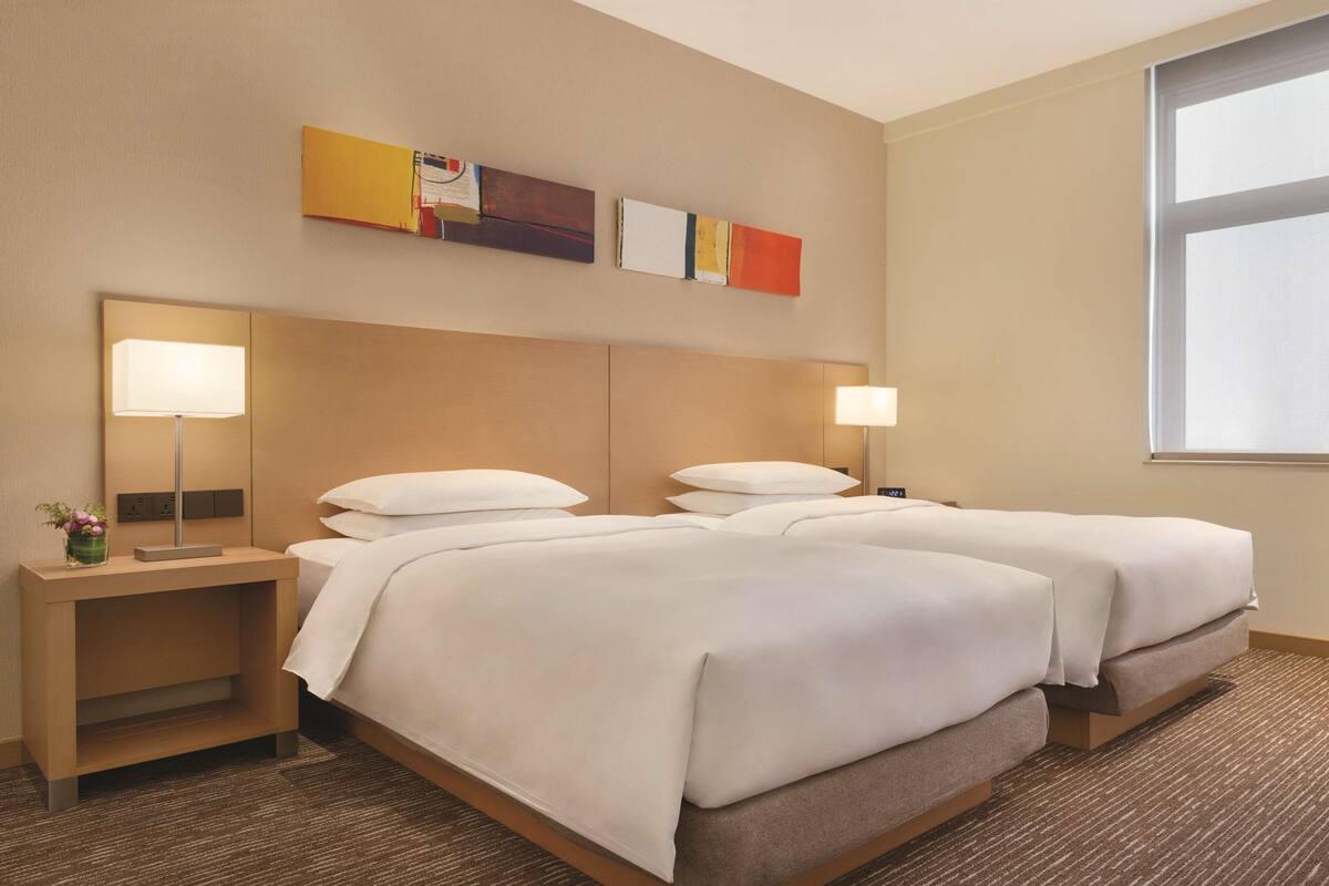 A hotel room with two beds, a nightstand with a lamp, and a window with blinds.