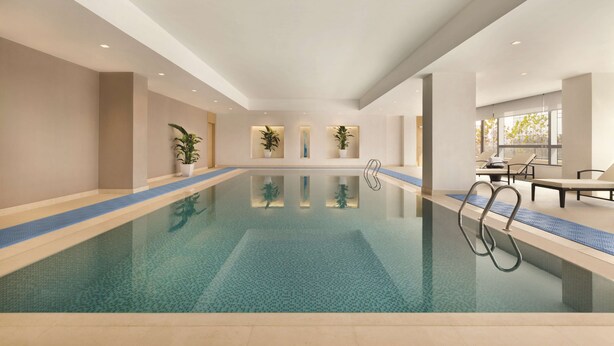 A large indoor swimming pool with clear blue water, surrounded by beige walls and a tiled floor. There are potted plants along the pool's edge and a seating area with chairs and a table on the right side. - Hyatt Place Beijing DaXing (Beijing)