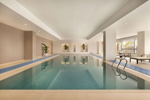 A large indoor swimming pool with clear blue water, surrounded by beige walls and a tiled floor. There are potted plants along the pool's edge and a seating area with chairs and a table on the right side. - Hyatt Place Beijing DaXing (Beijing)