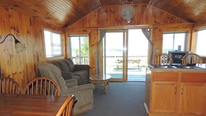 A cabin interior with a wooden dining table, a sofa, a kitchen area with a stove, and large windows.