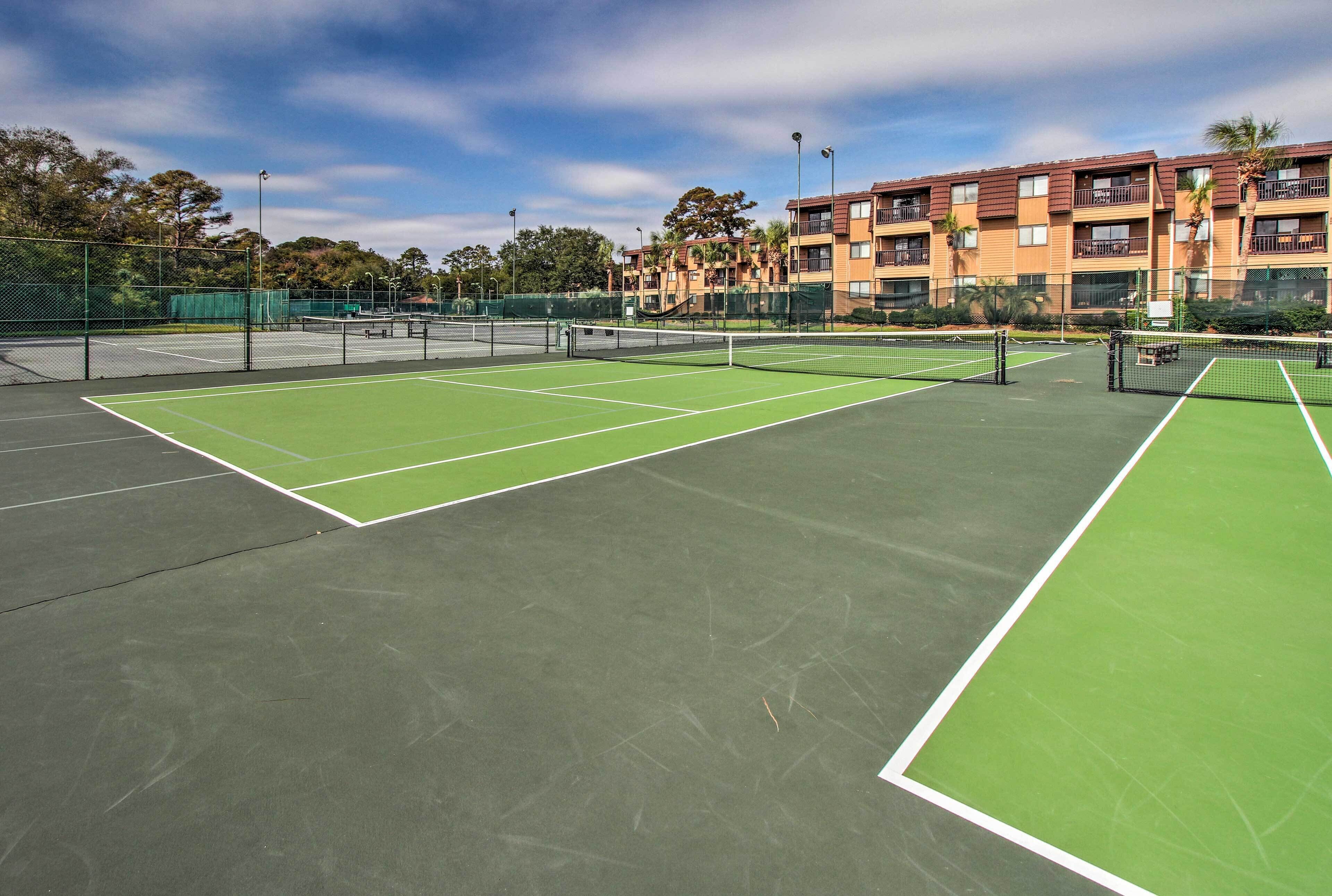A tennis court with green and black surfaces, surrounded by a fence, and adjacent to a multi-story building with balconies and trees.