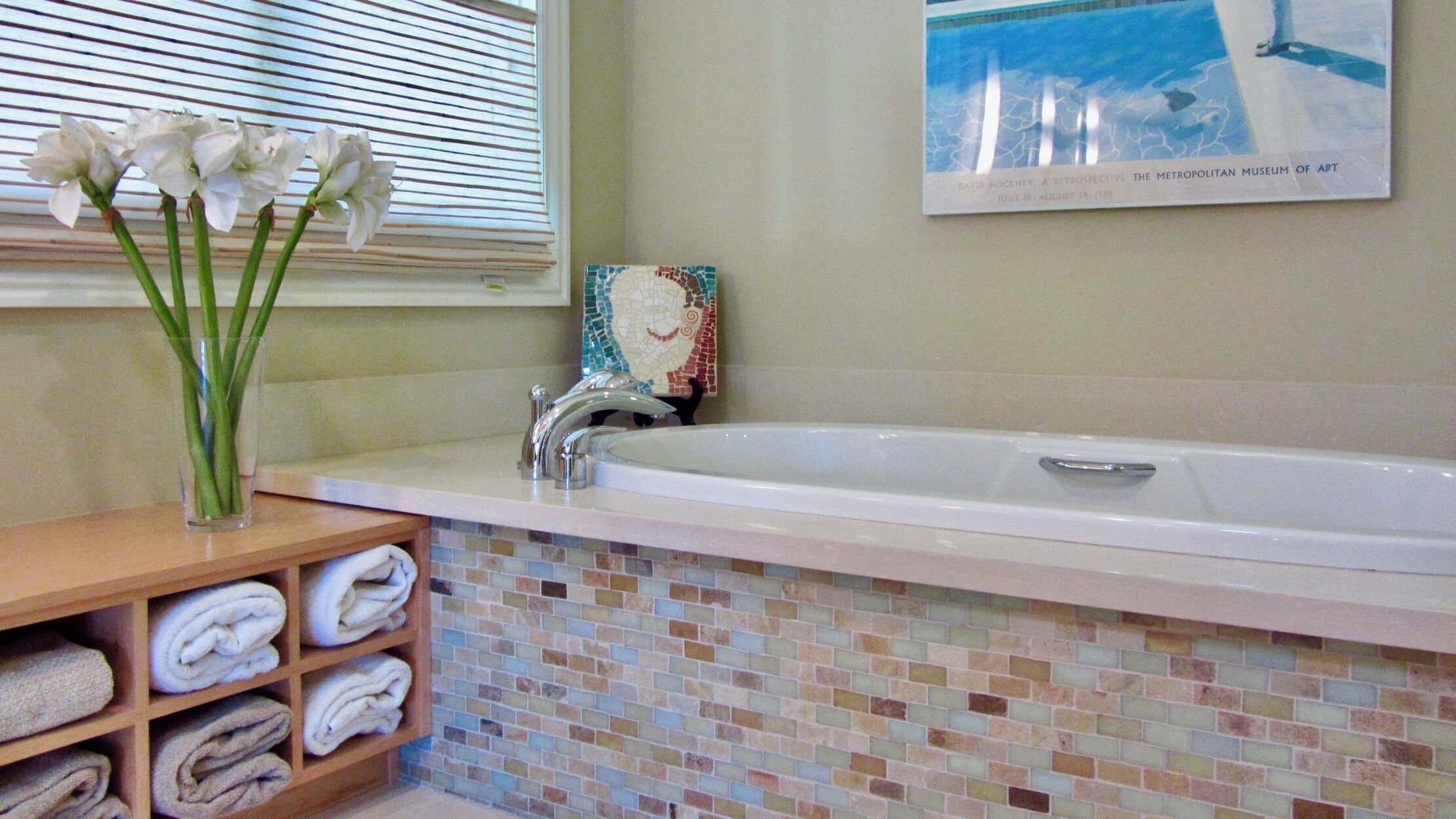 Une salle de bain avec un mur recouvert de mosaïques, une baignoire blanche, un vase en verre rempli de fleurs et un tableau encadré accroché au mur.