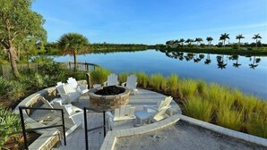 A circular outdoor seating area with white Adirondack chairs and a stone fire pit, surrounded by a paved walkway and a view of a calm lake with palm trees in the background. - Waterfront on Anna Maria Sound ~ Beaches just minutes away! (Bradenton)