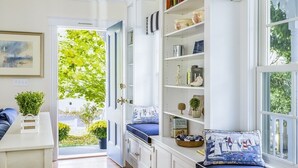 A room with a built-in bench with cushions, a bookshelf with various decorative items, and a view of greenery outside. - Spend your quintessential Cape Cod vacation, in a beautifully renovated 1862 Farm House on Stage Harbor Road! (Chatham)
