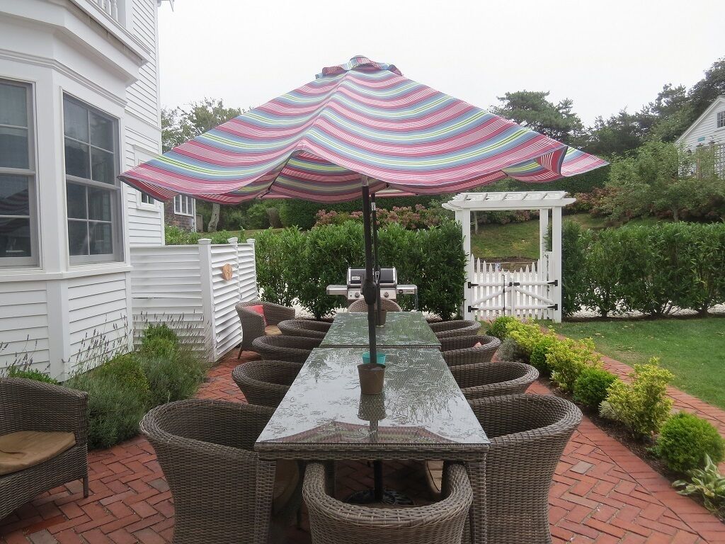 A patio with a long table and wicker chairs, a striped umbrella, and a white gazebo in the background.