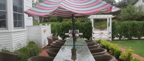 A patio with a long table and wicker chairs, a striped umbrella, and a white gazebo in the background.