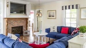 A living room with a fireplace, blue sofas, a white table, and a hanging light fixture. - Spend your quintessential Cape Cod vacation, in a beautifully renovated 1862 Farm House on Stage Harbor Road! (Chatham)