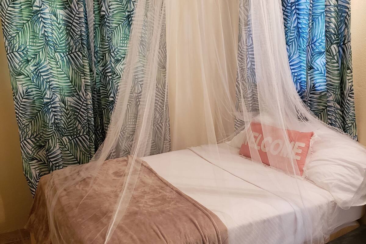 A bed with a canopy, a pink pillow with "WELCOME" text, and patterned curtains.