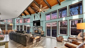 A spacious living room with a stone fireplace, large windows, and a ceiling fan. - Amazing Lake Views, Game room, Private Beach, Hot Tub, Boats and Firepit (Tobyhanna)