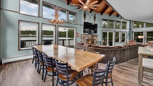 A spacious dining area with a long wooden table and chairs, a stone fireplace, and a large TV mounted on the wall. - Amazing Lake Views, Game room, Private Beach, Hot Tub, Boats and Firepit (Tobyhanna)