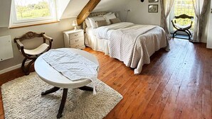Een slaapkamer met een schuine kap, een bed, een stoel, een klein tafeltje en een raam met gordijnen.