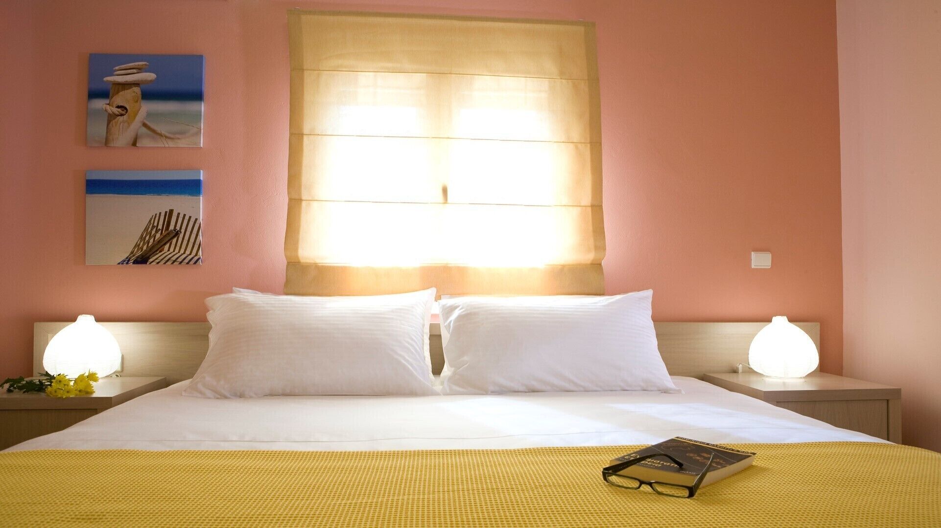 A hotel room with a bed, two bedside lamps, a window with blinds, and two wall art pieces.