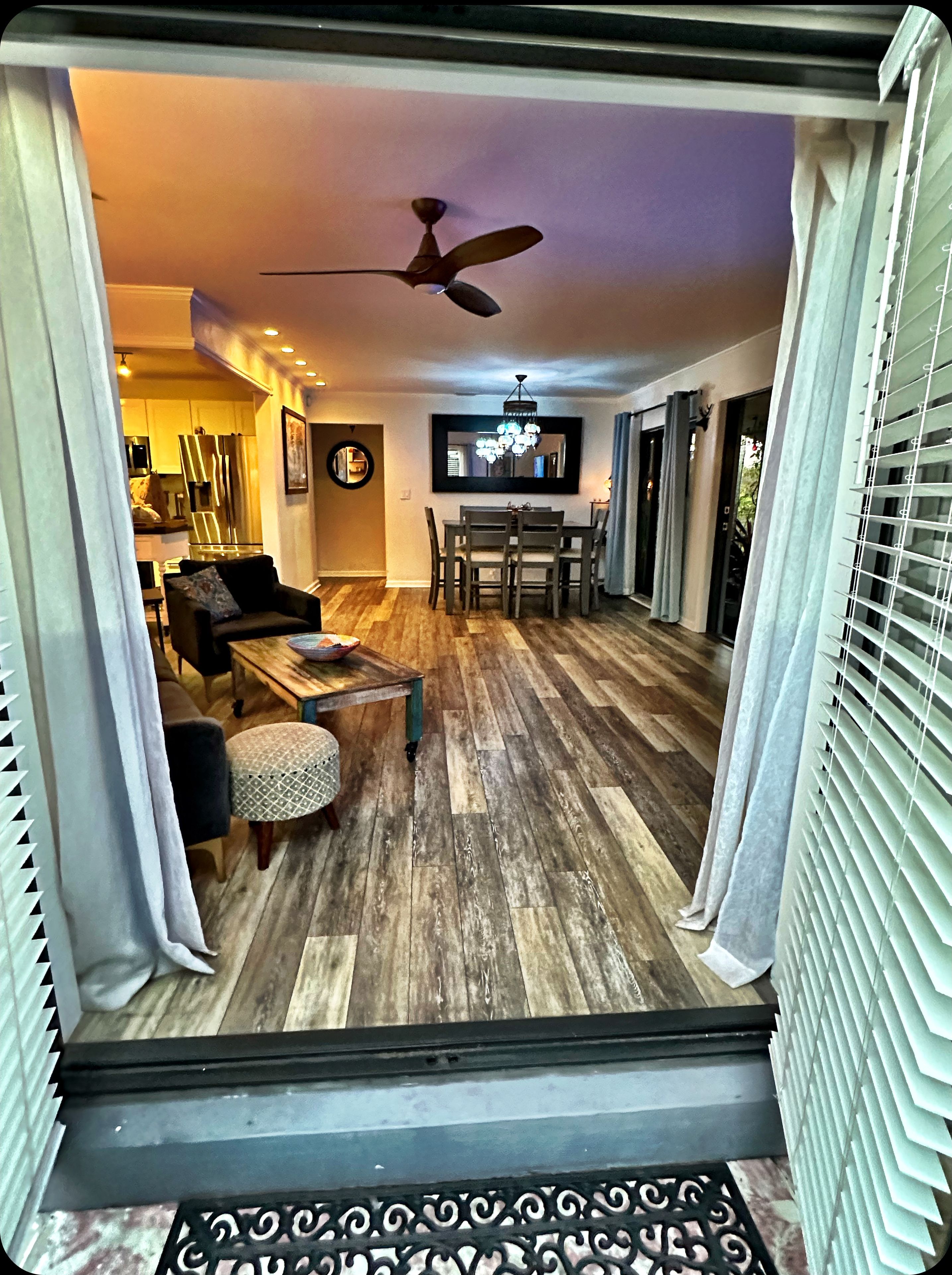 A spacious living area with a wooden floor, a ceiling fan, a dining table with chairs, and a kitchen in the background.