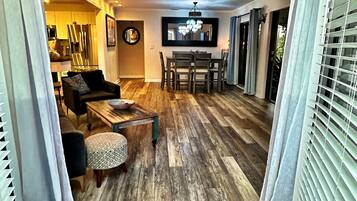 A spacious living area with a wooden floor, a ceiling fan, a dining table with chairs, and a kitchen in the background.