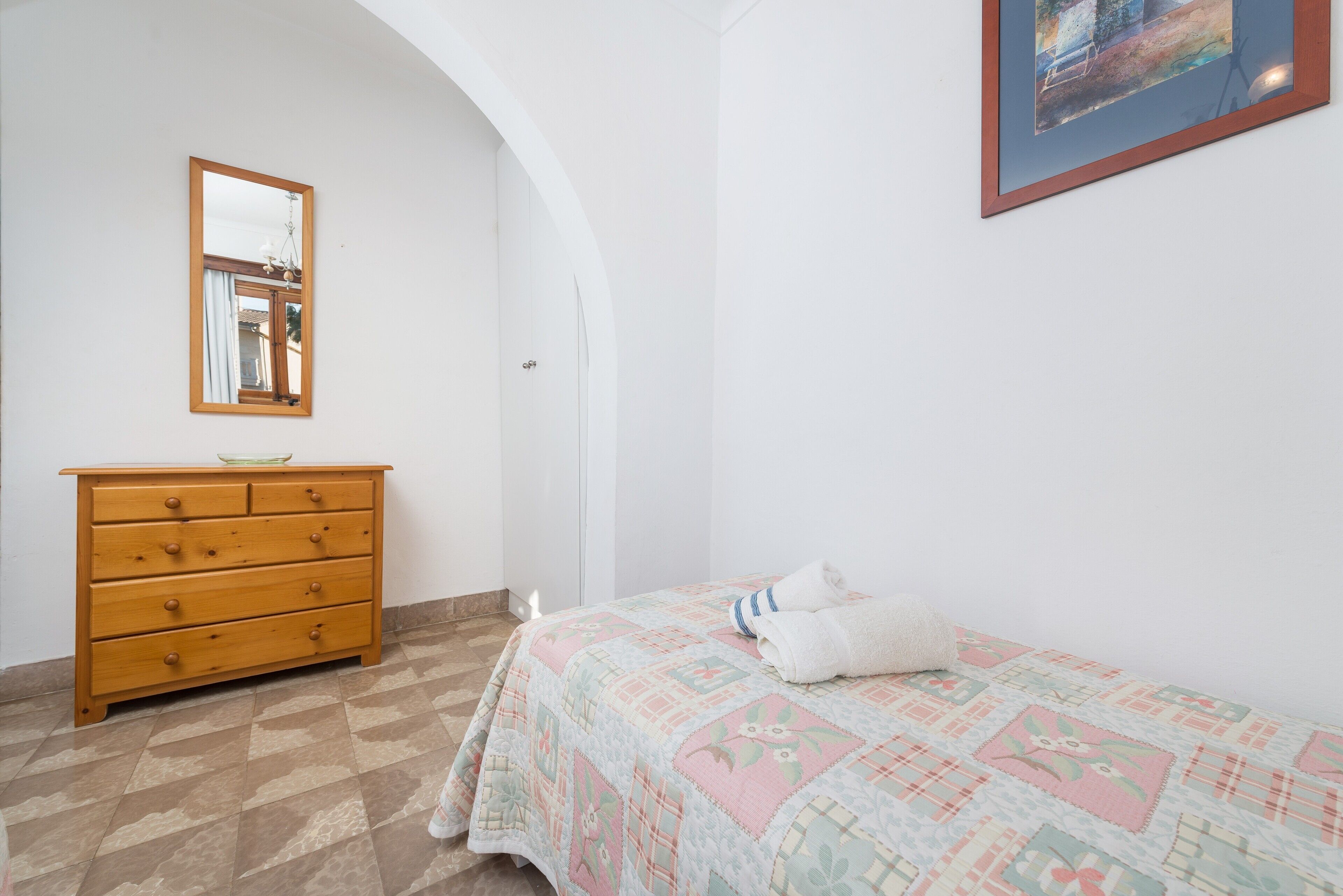 A bedroom with a wooden dresser, a bed with a quilt, and a painting on the wall.