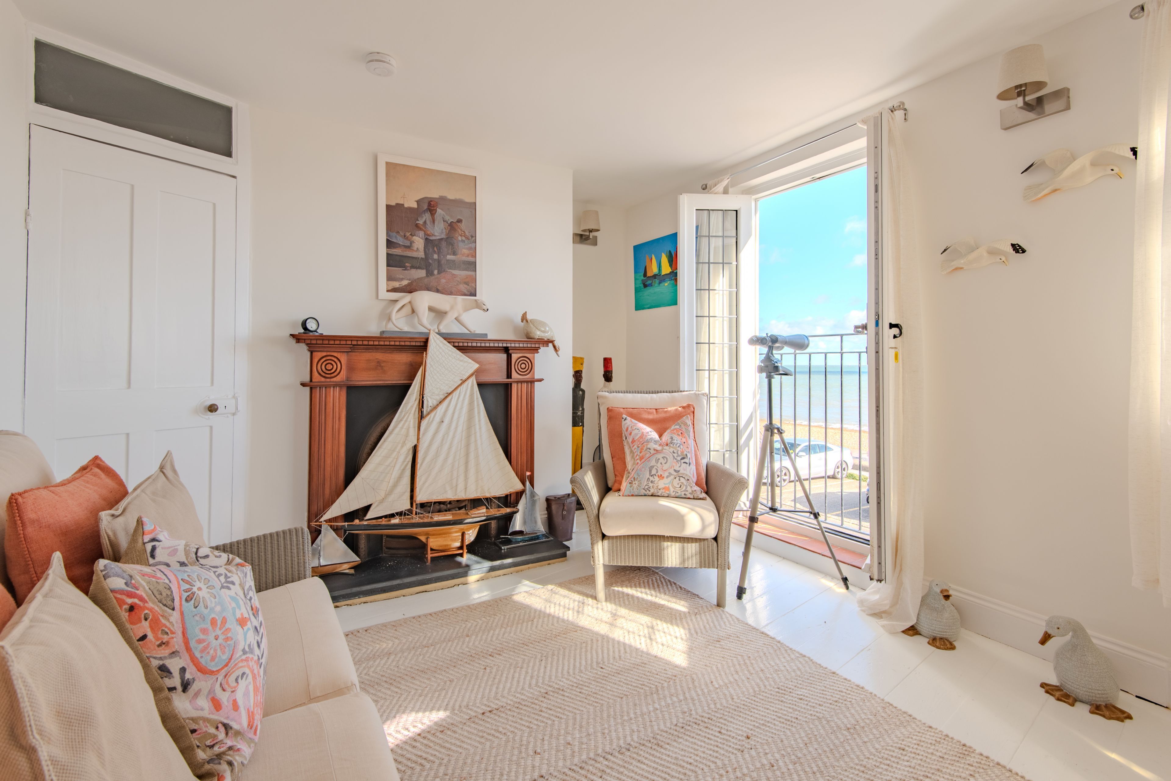 A living room with a fireplace, a sailboat model, a chair, and a view of the sea through a balcony door.