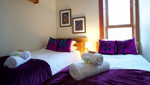 A hotel room with two beds, purple bedding, a wooden shelf, and two framed pictures on the wall. - Castle View Vintage Retreat (Edinburgh)
