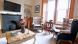 A living room with a fireplace, a television mounted above it, a plaid armchair, a small round table, and a large window with natural light. - Castle View Vintage Retreat (Edinburgh)