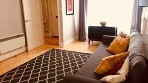Un salon chaleureux avec un canapé, un tapis à motifs, une petite table sur laquelle se trouve un bol, et une porte qui donne sur une autre pièce.