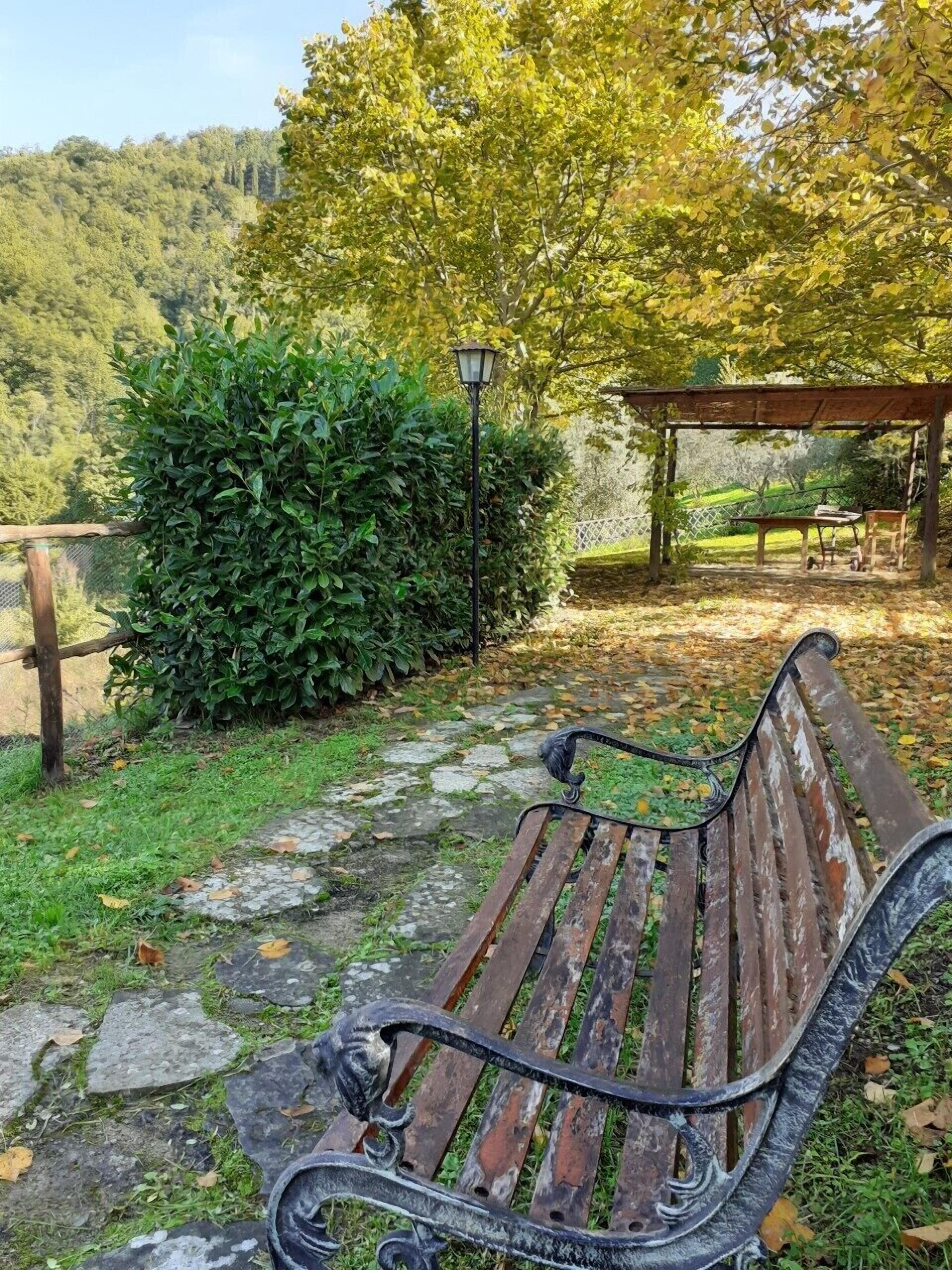 Un banc en bois avec des accoudoirs en métal, un chemin pavé de pierres, un lampadaire et une pergola en bois avec une table et des chaises.