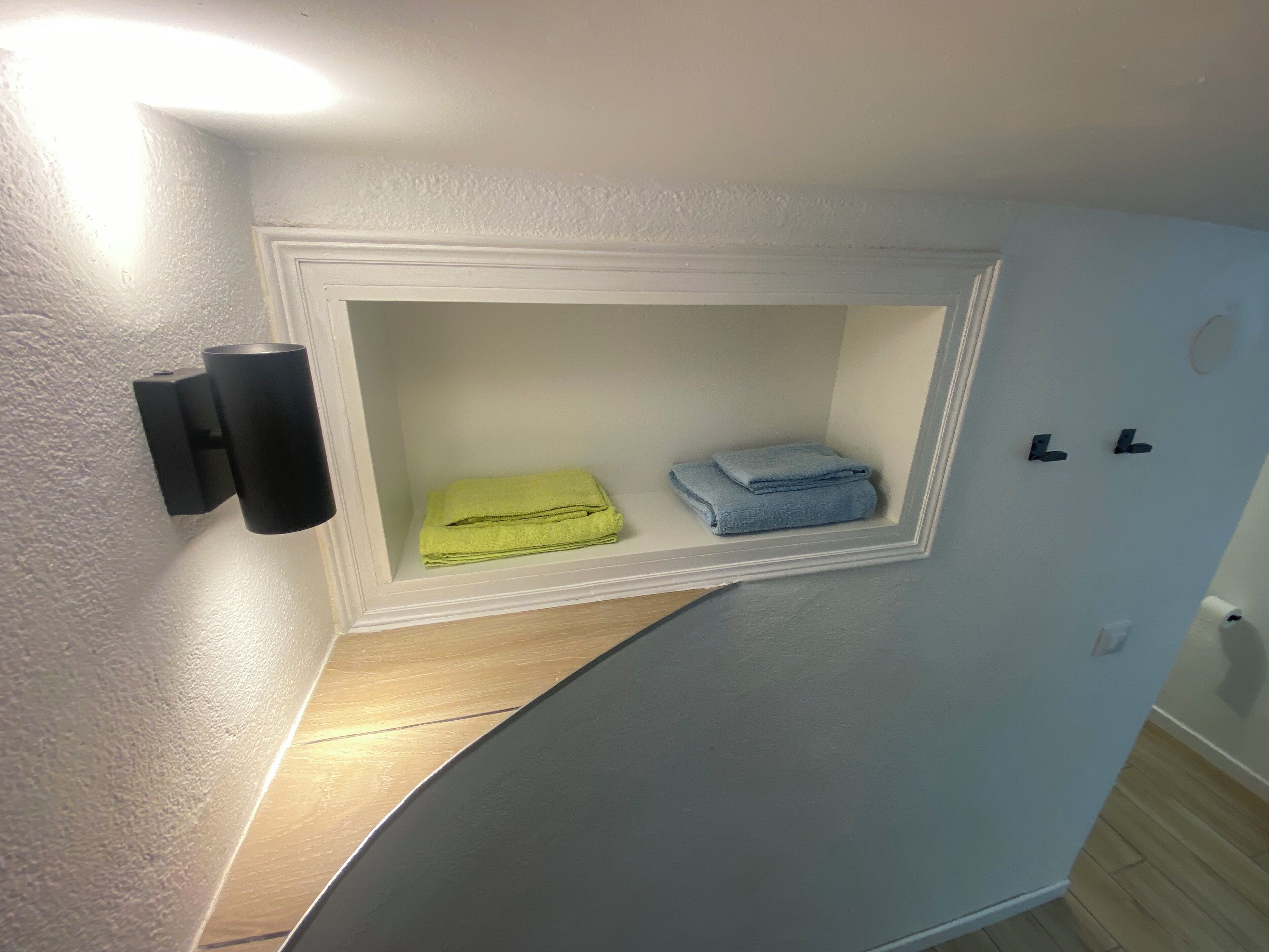 Une salle de bain avec une étagère intégrée contenant deux serviettes pliées, un luminaire fixé au mur et une poignée de porte.