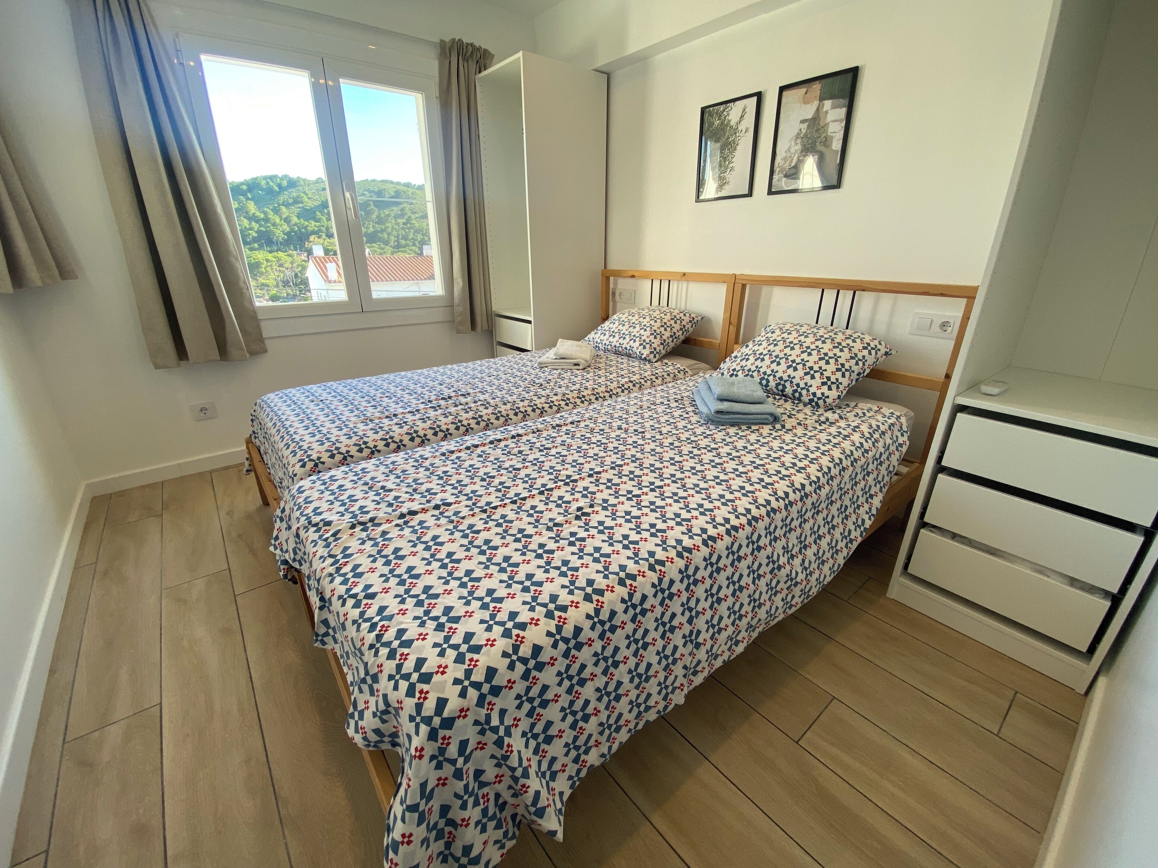 A bedroom with two beds, a wooden floor, a window with curtains, and a small desk with drawers.