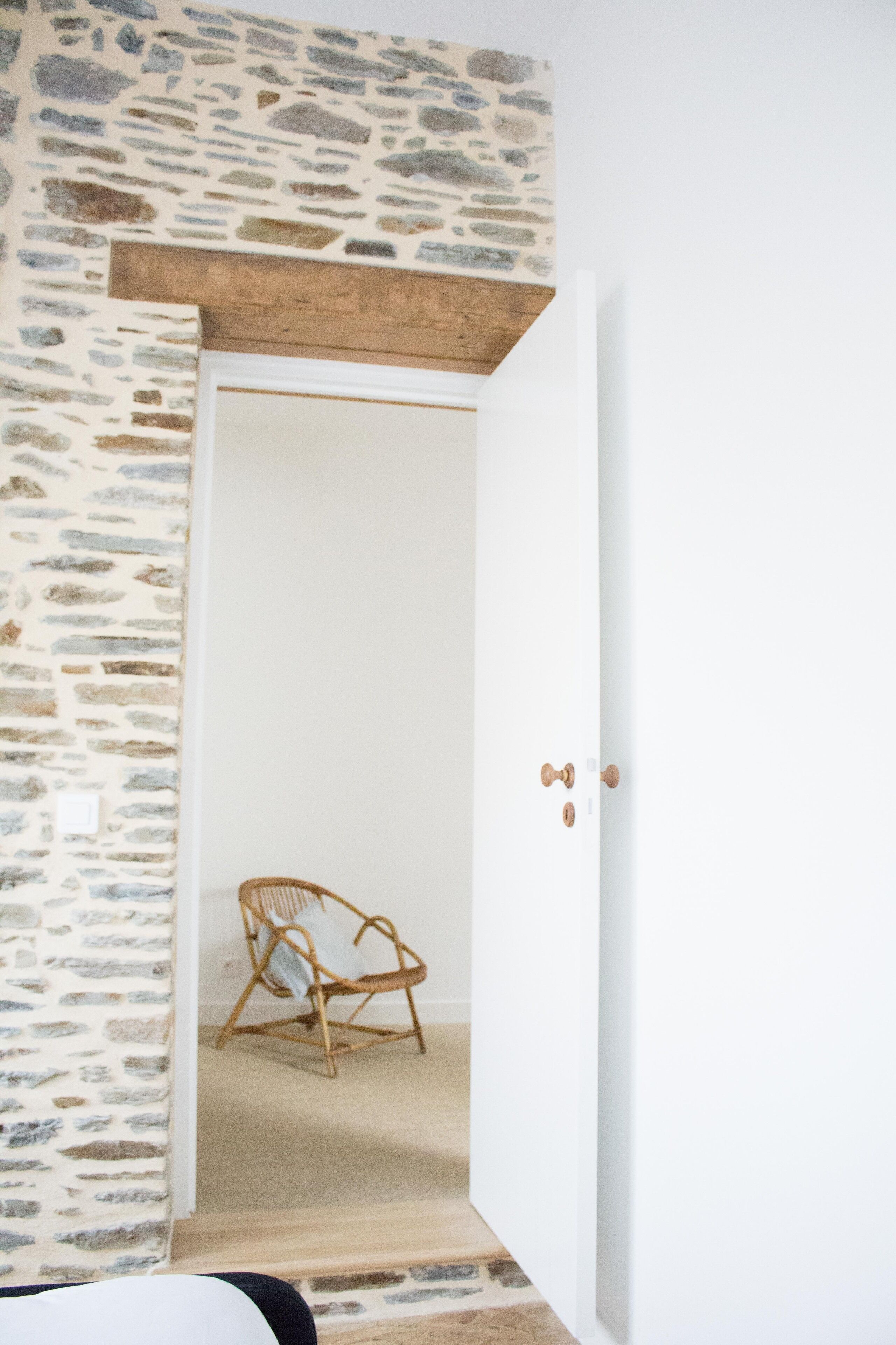 A room with a stone wall, a wooden ceiling, a white door, and a wicker chair.