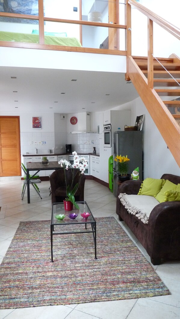 Amplia sala de estar con escalera, una mesa de comedor con sillas, un sofá con cojines y una cocina al fondo.