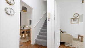 A hallway with wooden flooring, a staircase, a dining area with a table and chairs, and a living area with a sofa and a small table.