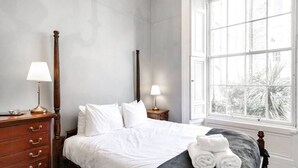 A bedroom with a four-poster bed, two bedside tables with lamps, a dresser, and a large window with a view of trees.