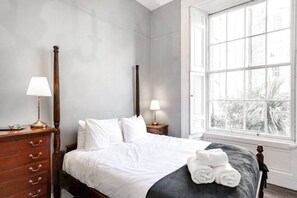 A bedroom with a four-poster bed, two bedside tables with lamps, a dresser, and a large window with a view of trees.