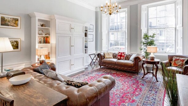 A spacious living room with a leather sofa, a patterned rug, and a large window with a view of buildings.